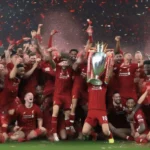 Liverpool celebrating their Premier League title win in the 2019-2020 season. The players are holding the trophy aloft and are surrounded by confetti and fireworks.